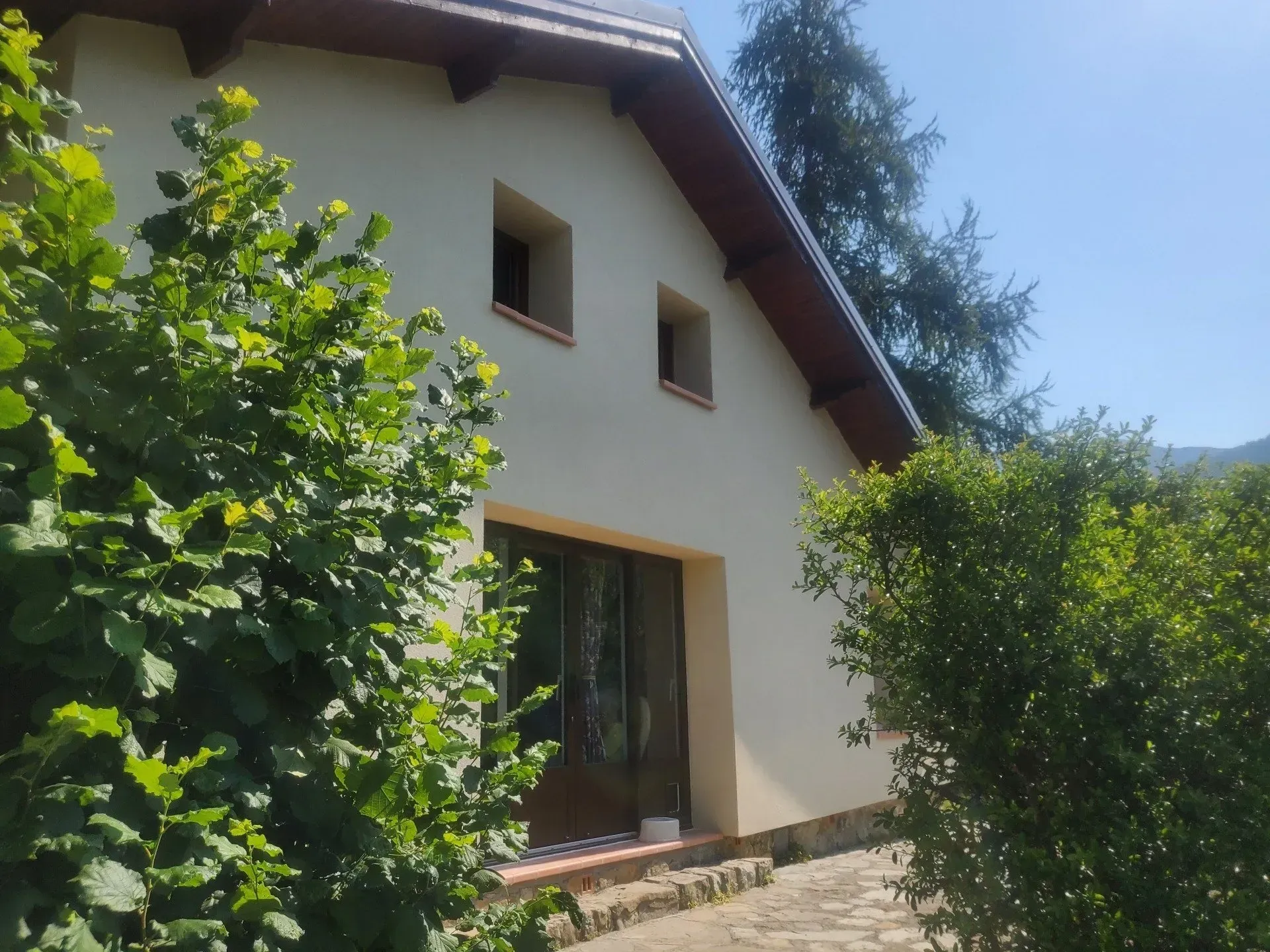 Maison beige avec une porte marron, deux petites fenêtres et des buissons verts devant.