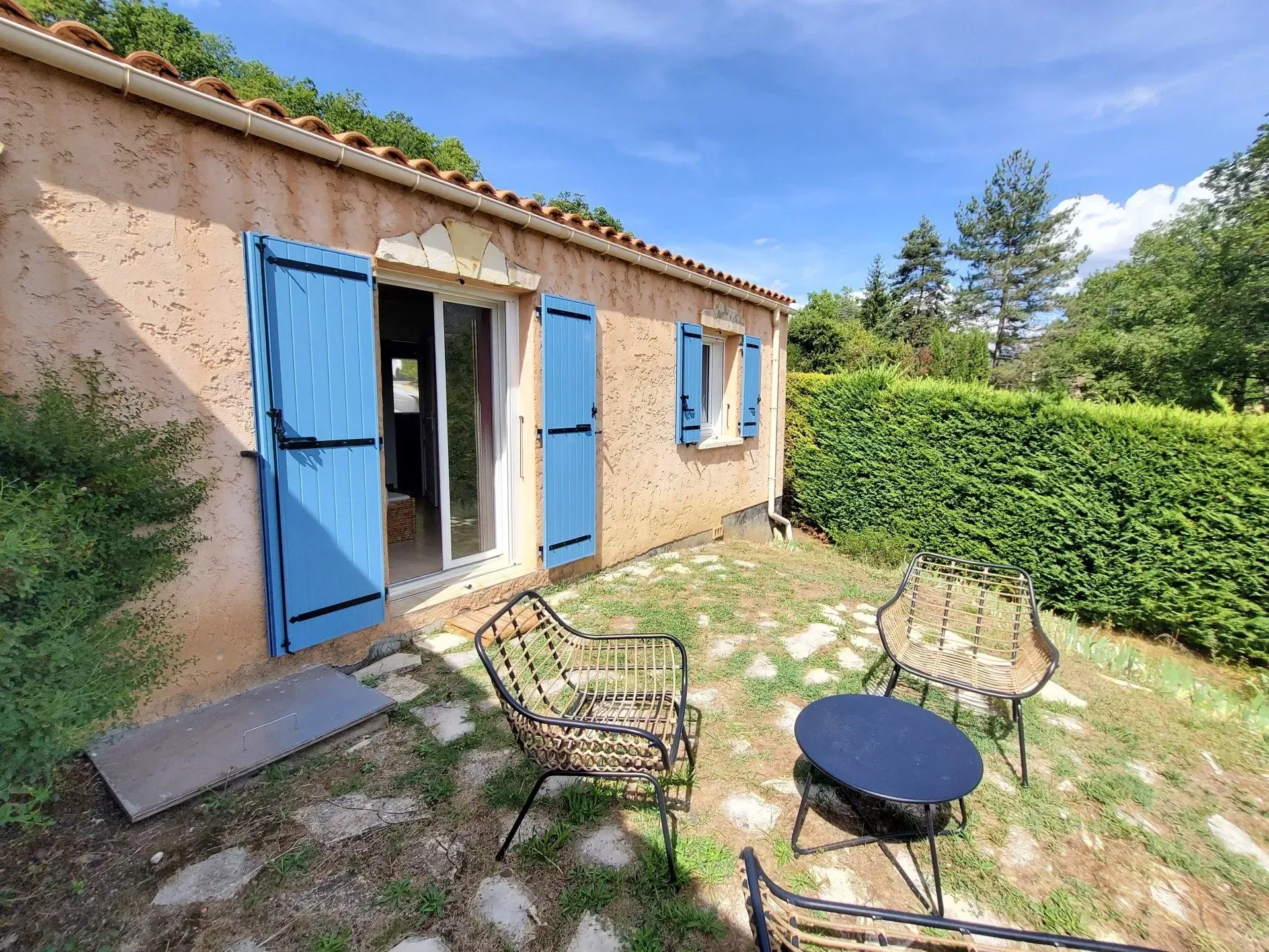 Petit cottage avec volets bleus, mobilier de jardin et haie, par une journée ensoleillée.