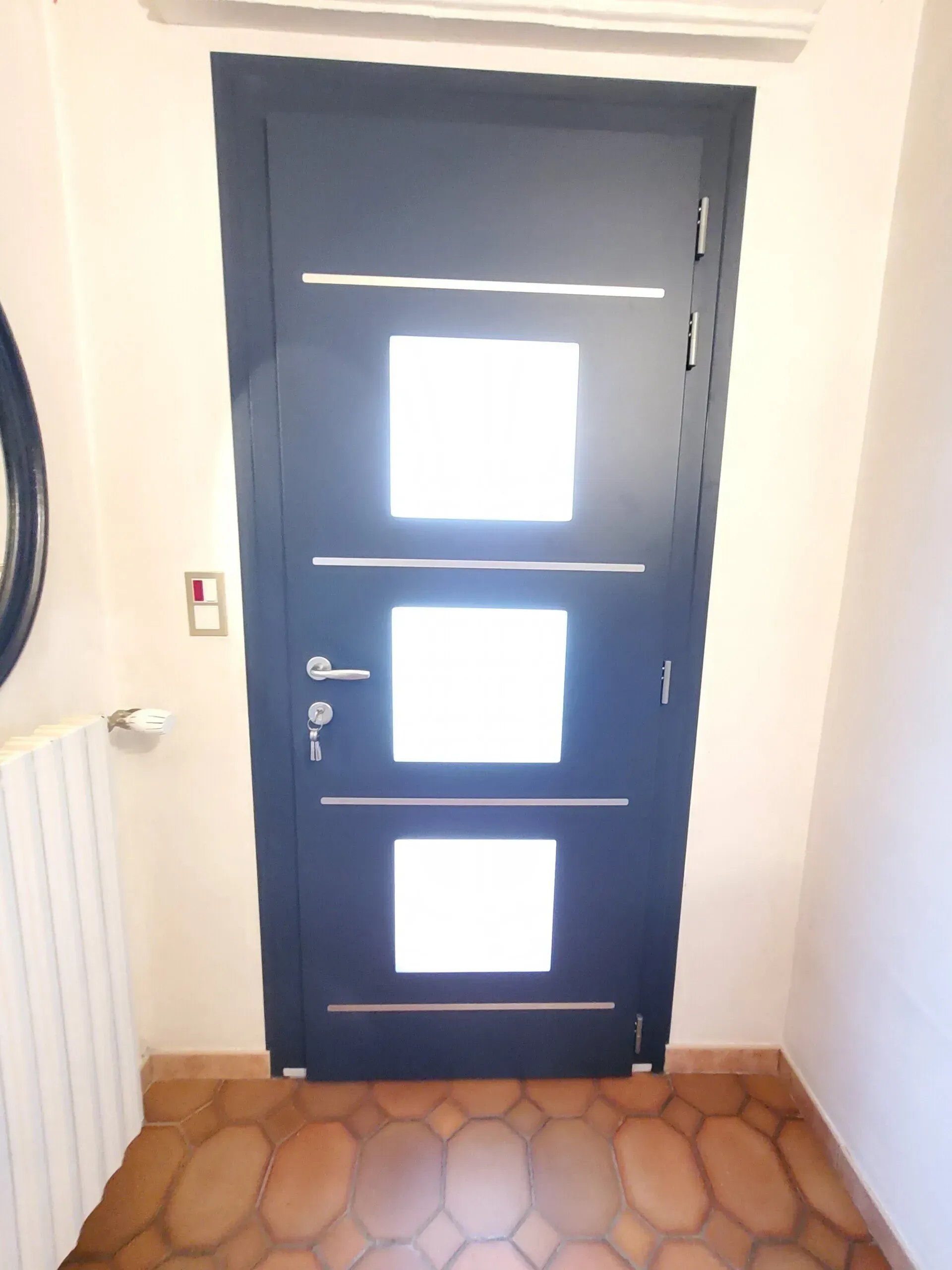 Porte d'entrée noire avec trois panneaux de verre carrés, quincaillerie argentée et accents argentés horizontaux.