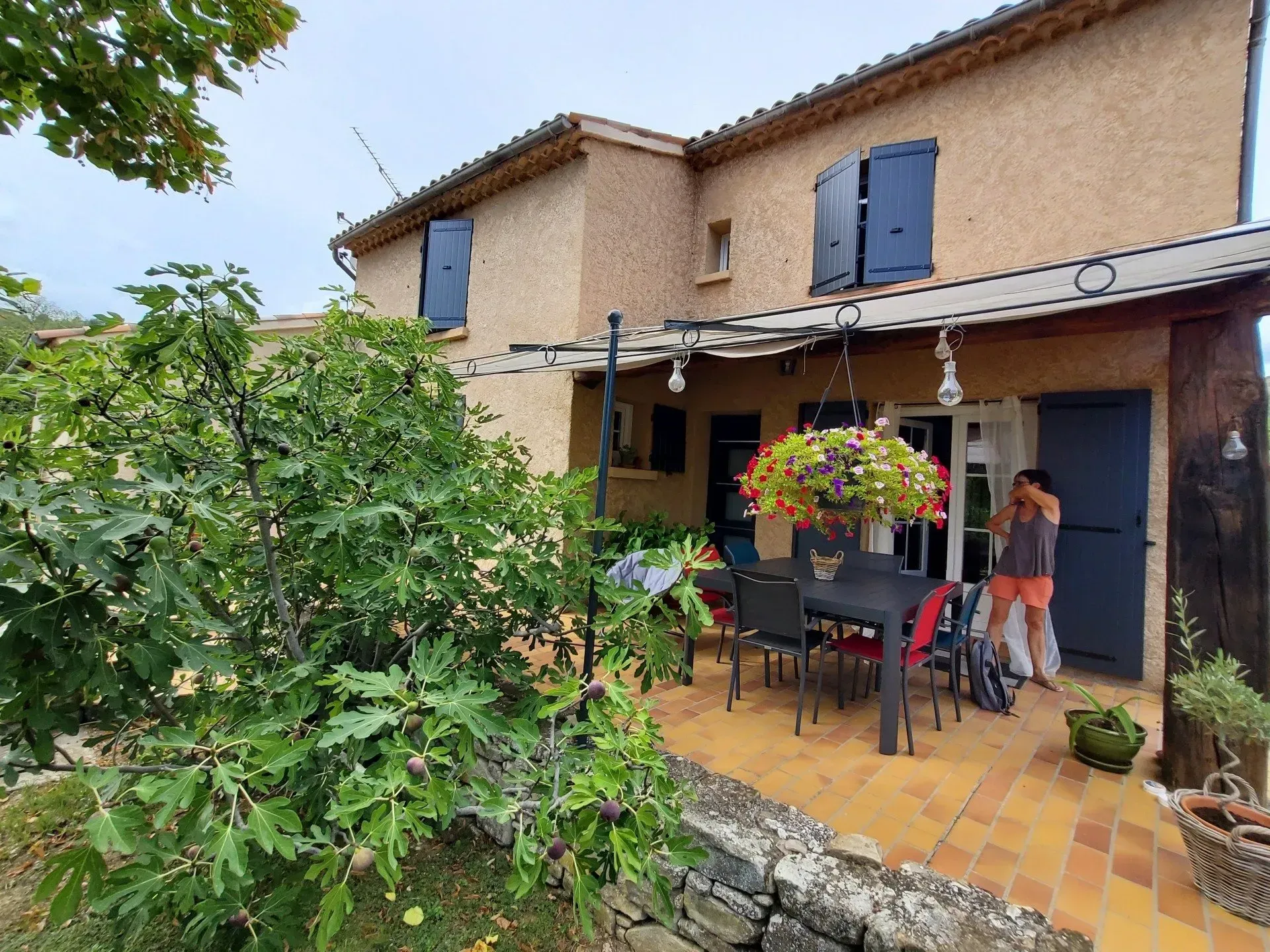 Maison avec patio en terre cuite, ensemble de salle à manger d'extérieur et panier de fleurs suspendu ; deux personnes près d'une porte.