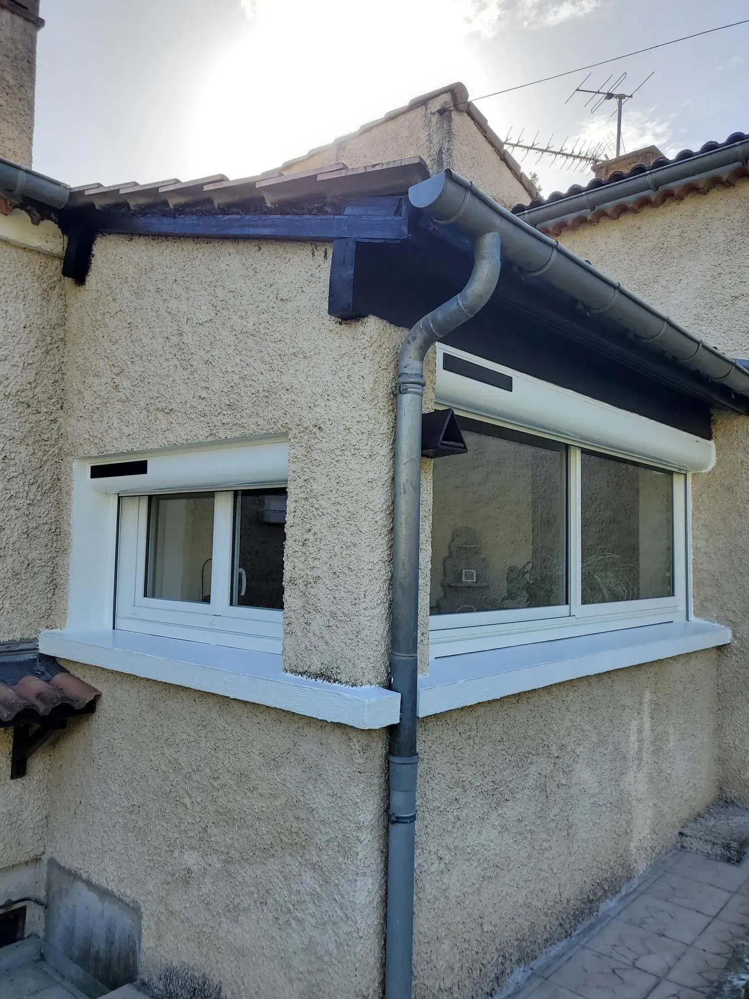 Angle extérieur d'un bâtiment avec des fenêtres blanches, un toit sombre et une gouttière grise.