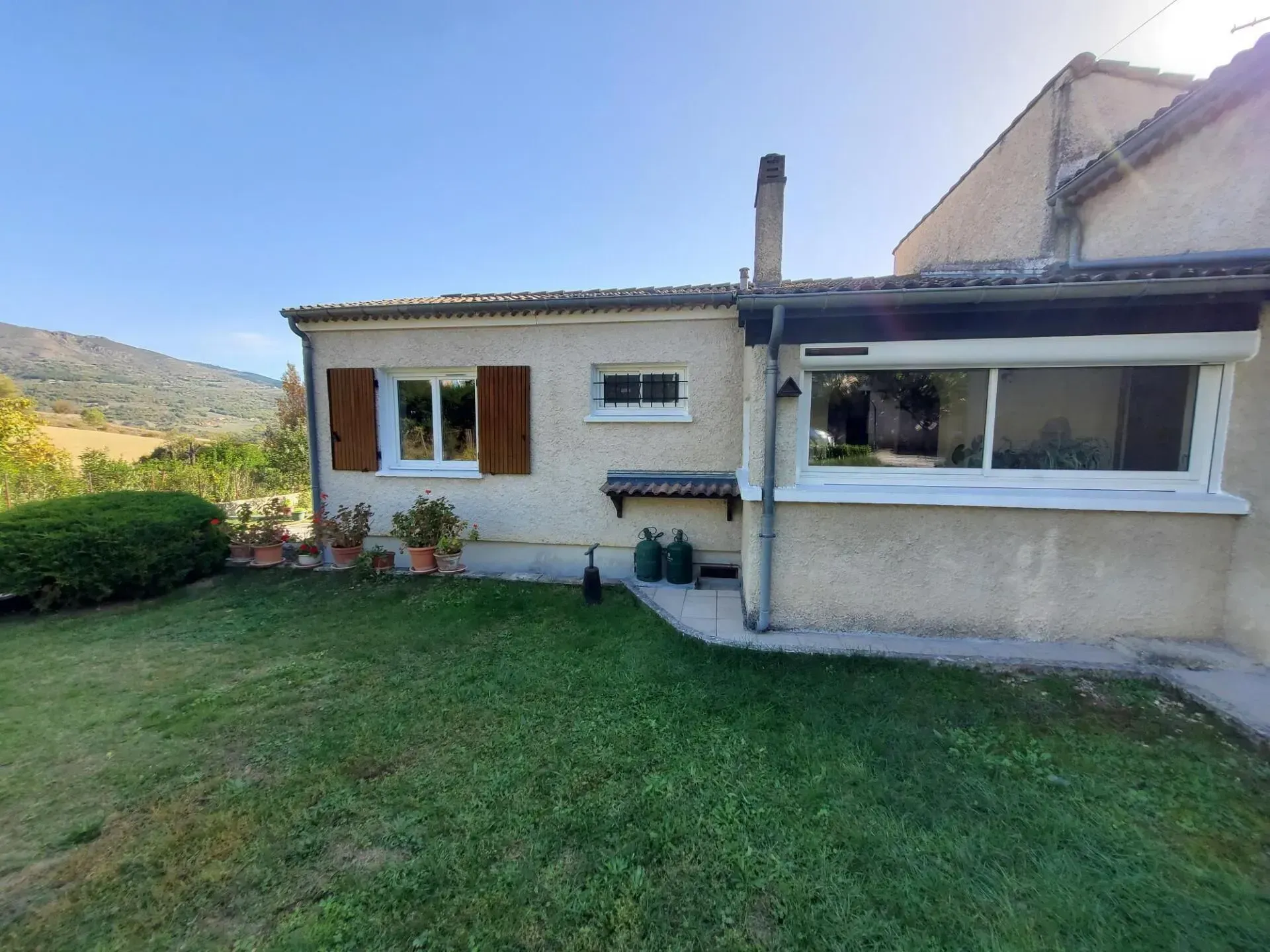 Maison aux volets marrons, pelouse verte, montagnes à l'horizon par une journée ensoleillée.