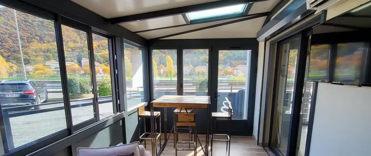 Véranda avec table et chaises, grandes fenêtres donnant sur les couleurs d'automne et un véhicule.