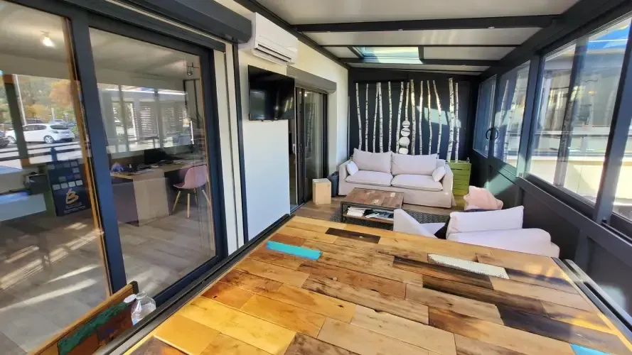 Véranda avec table en bois, canapé et parois vitrées ; bureau visible par une porte coulissante.