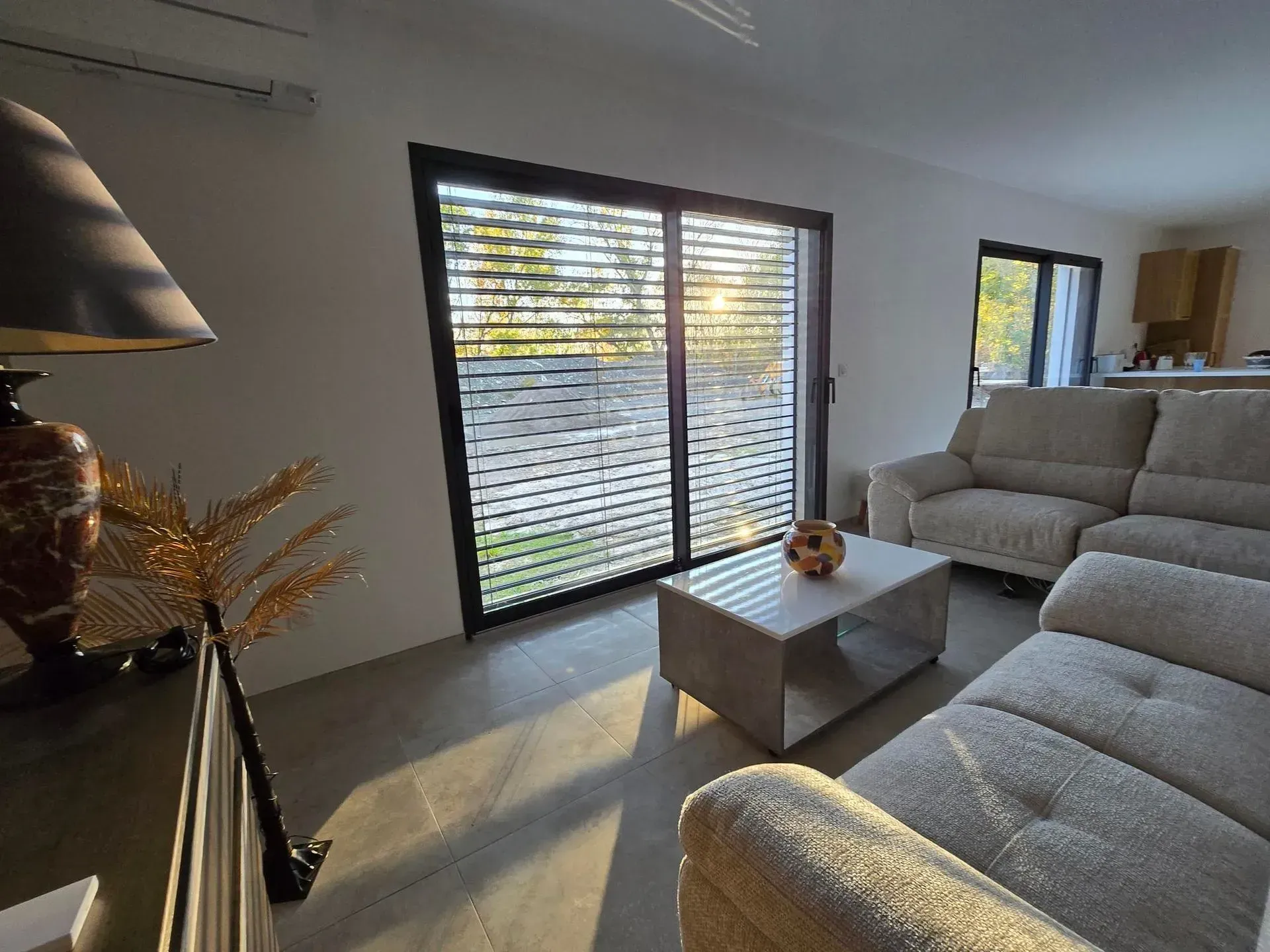 Salon avec une grande fenêtre recouverte de stores horizontaux, un canapé et une table basse.