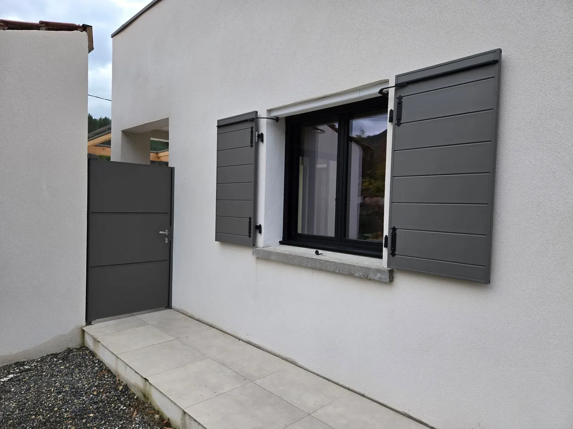 Volets gris sur un bâtiment blanc, avec une porte grise assortie et une allée en pierre.