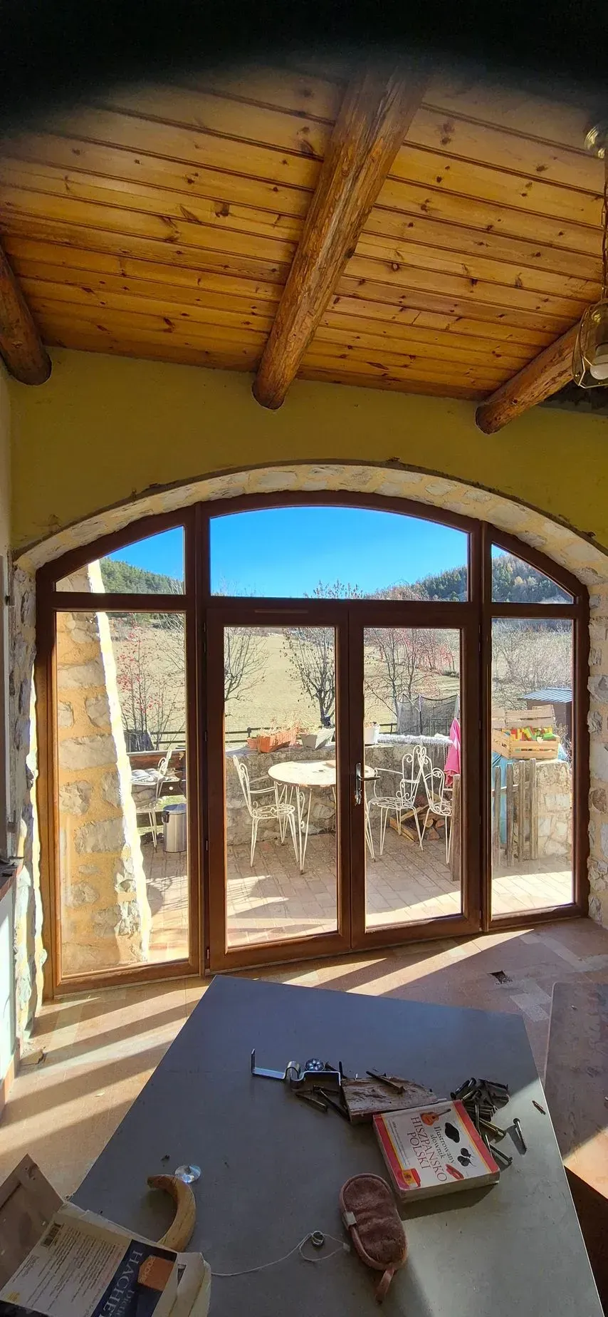 Vue intérieure avec une grande fenêtre cintrée, un plafond en bois aux poutres apparentes, une table au premier plan et une vue extérieure ensoleillée.