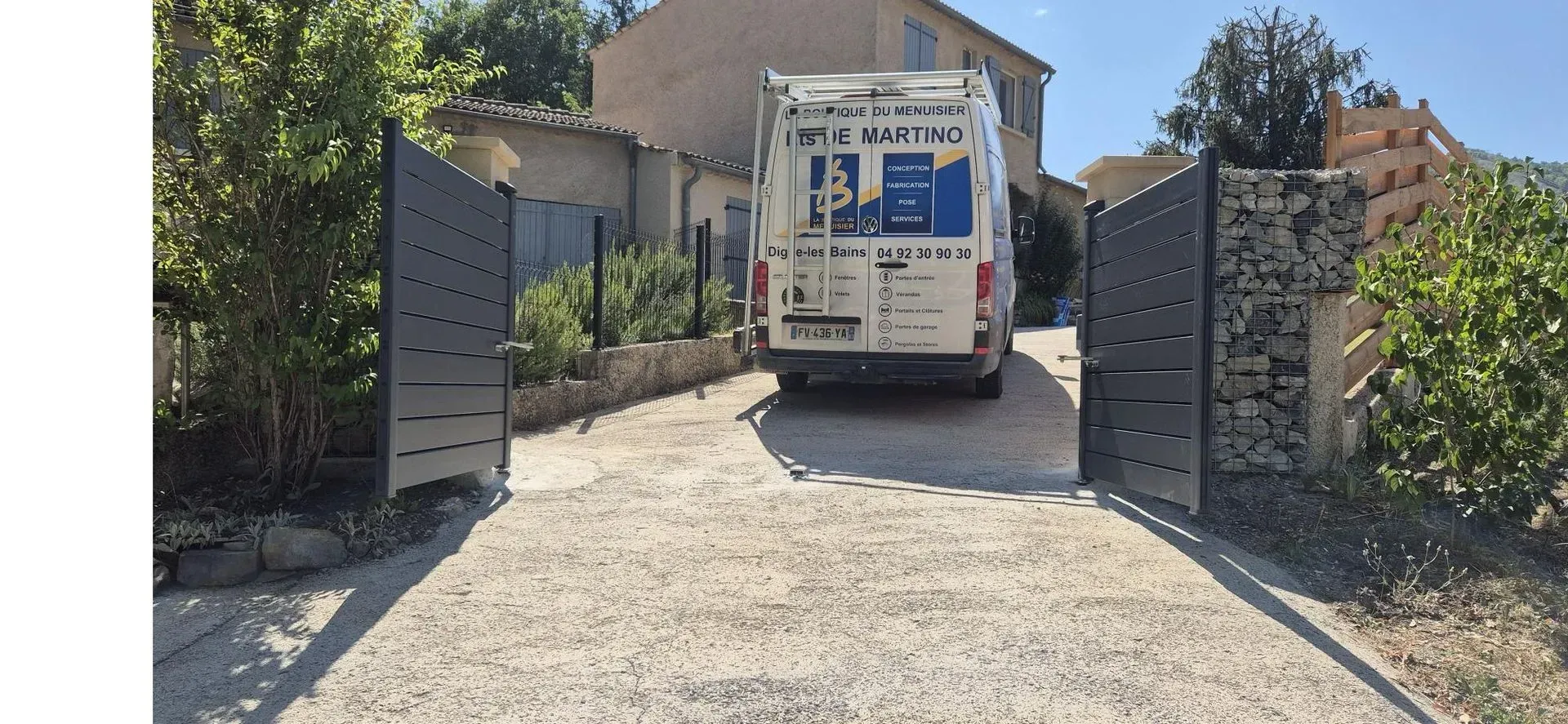 Portail noir ouvert, camionnette blanche garée sur une allée de gravier devant un bâtiment.