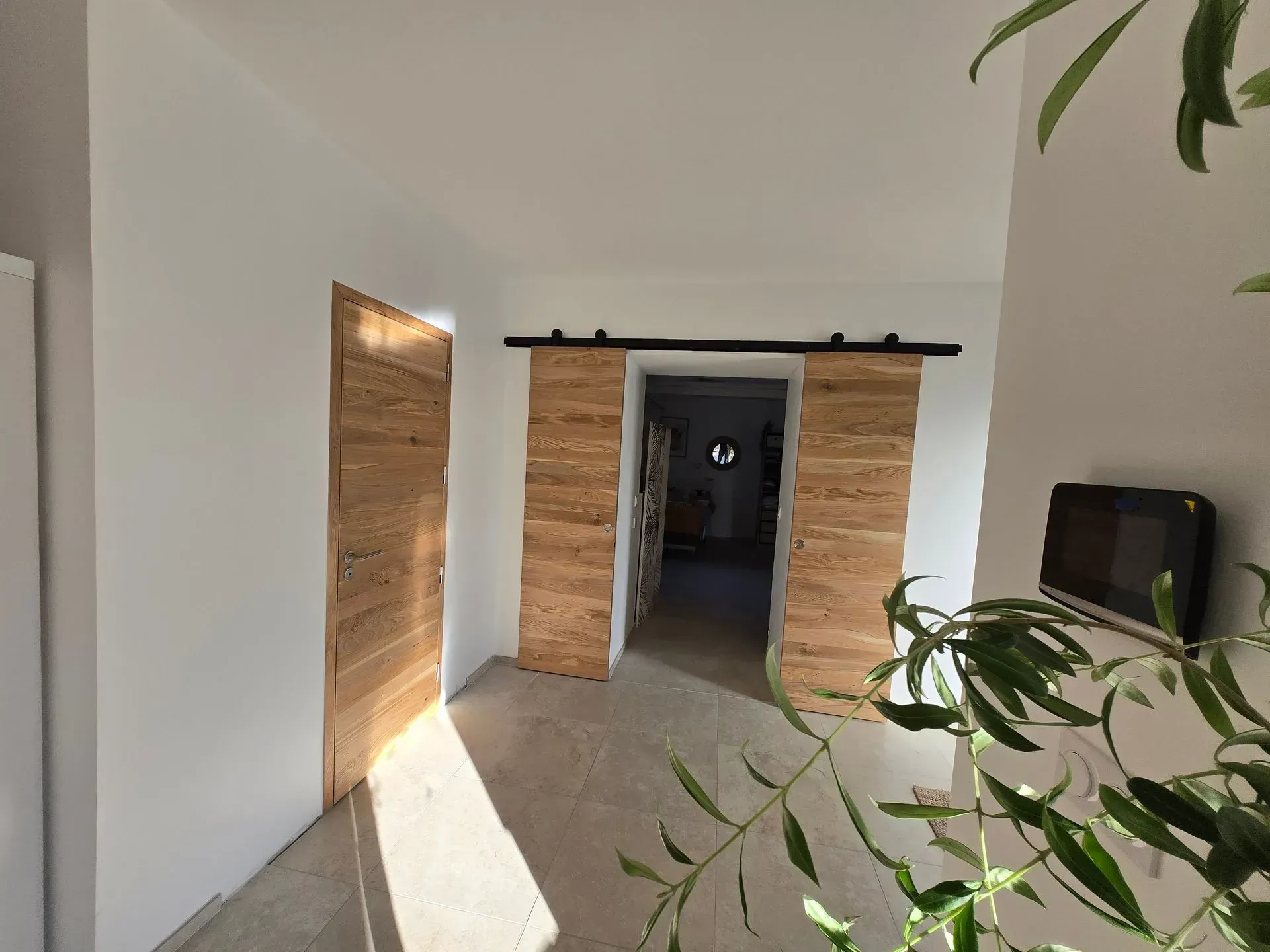 Couloir aux murs gris clair, sol en béton et portes en liège, l'une ouverte, l'autre fermée. Branches d'olivier à droite.