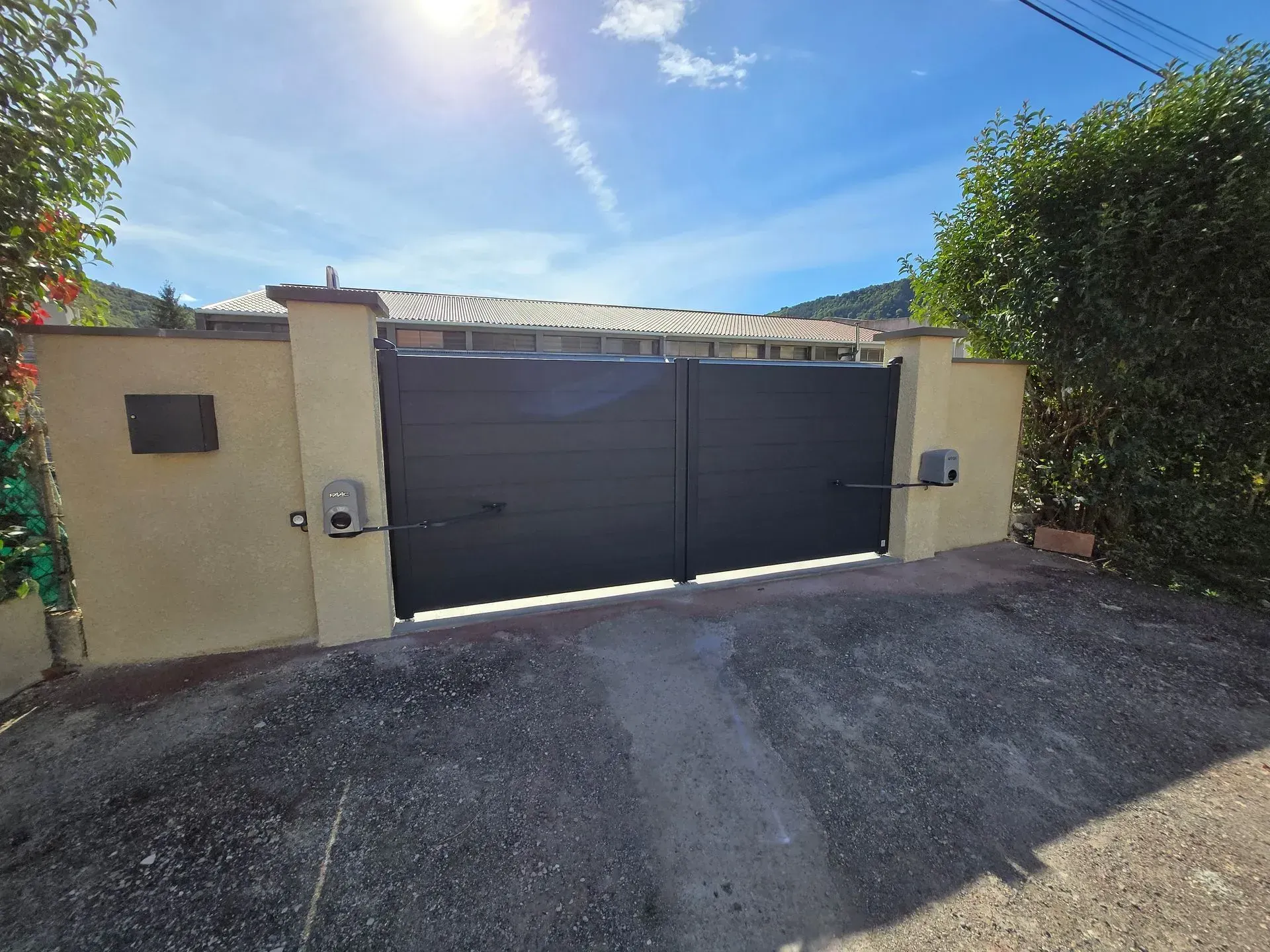 Portail en métal noir entre des piliers beiges, allée de gravier. Belle journée ensoleillée, ciel bleu.