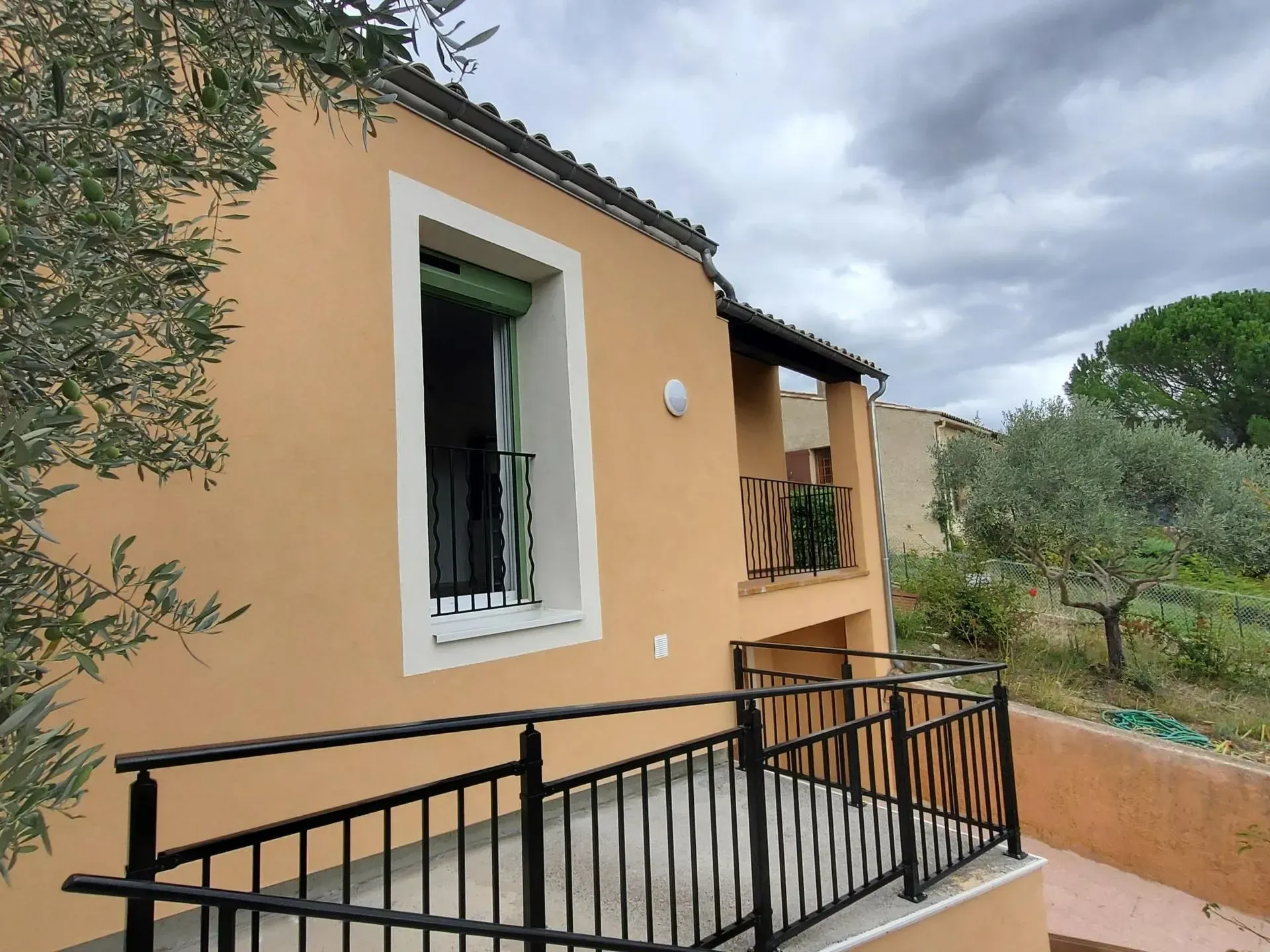 Vue extérieure d'une maison en stuc avec une balustrade, une fenêtre et un balcon noirs, sur fond de ciel nuageux.