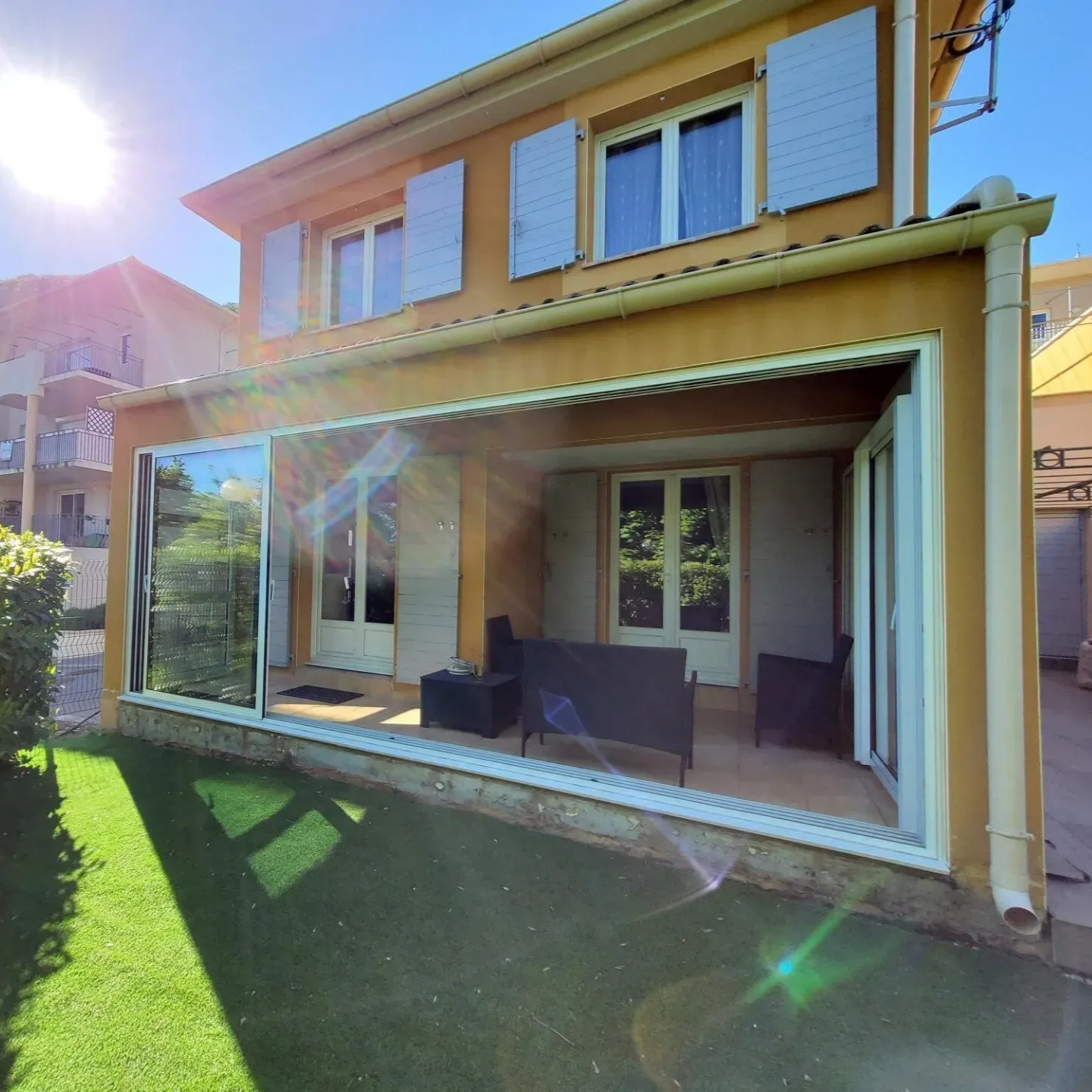 Maison jaune avec de grandes portes coulissantes en verre, patio ensoleillé, pelouse verte et vue sur la montagne.
