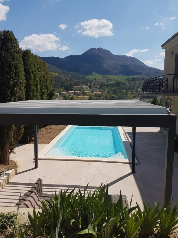 Piscine sous une pergola, avec les montagnes en toile de fond et un ciel bleu.