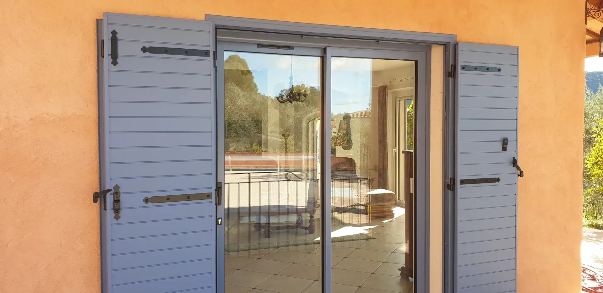 Des volets bleus encadrent une porte coulissante en verre sur un mur en stuc orange.