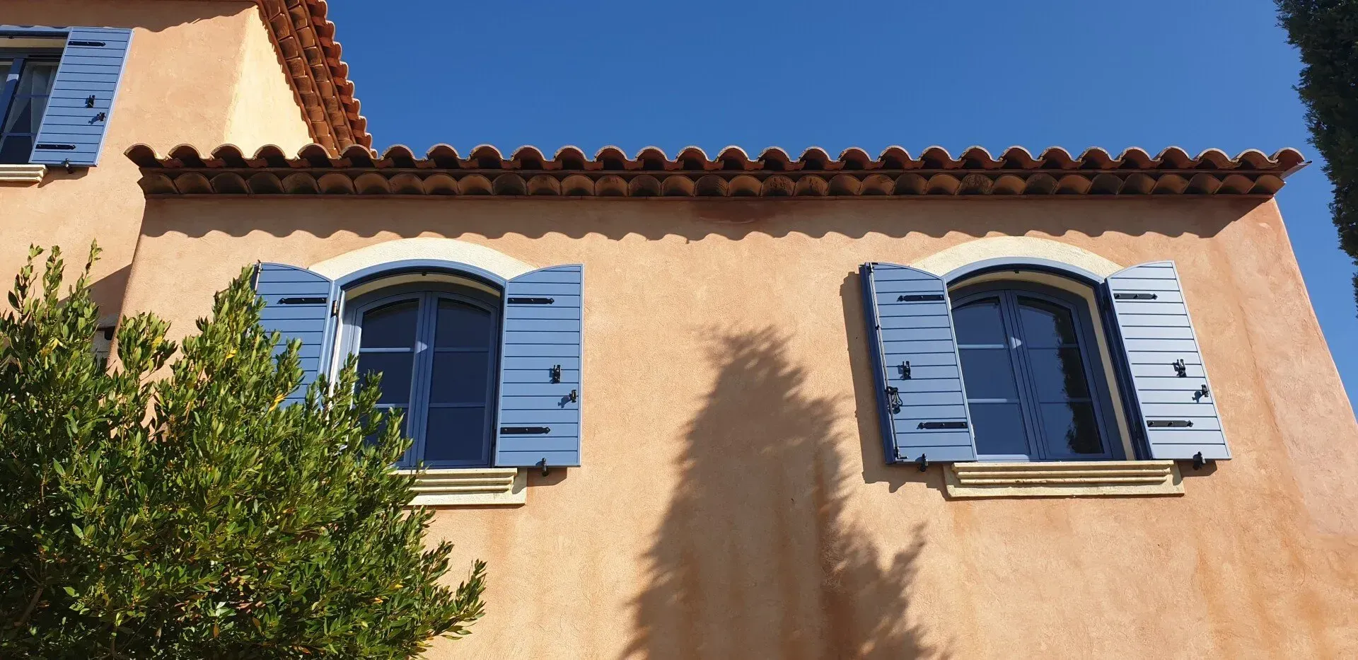 Façade d'un bâtiment avec des volets bleus sur des fenêtres cintrées, sous un toit de tuiles en terre cuite, sur fond de ciel bleu clair.