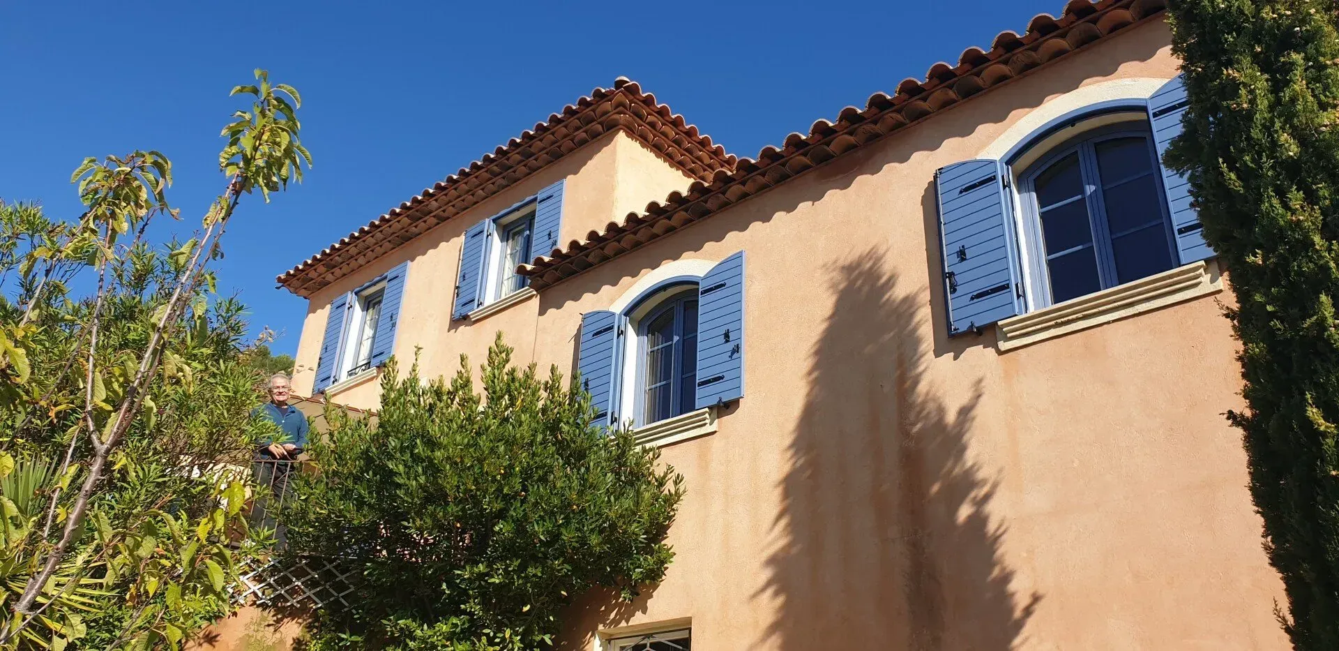 Un bâtiment couleur pêche, avec des volets bleus et un toit en tuiles de terre cuite, se détachant sur un ciel d'un bleu éclatant.