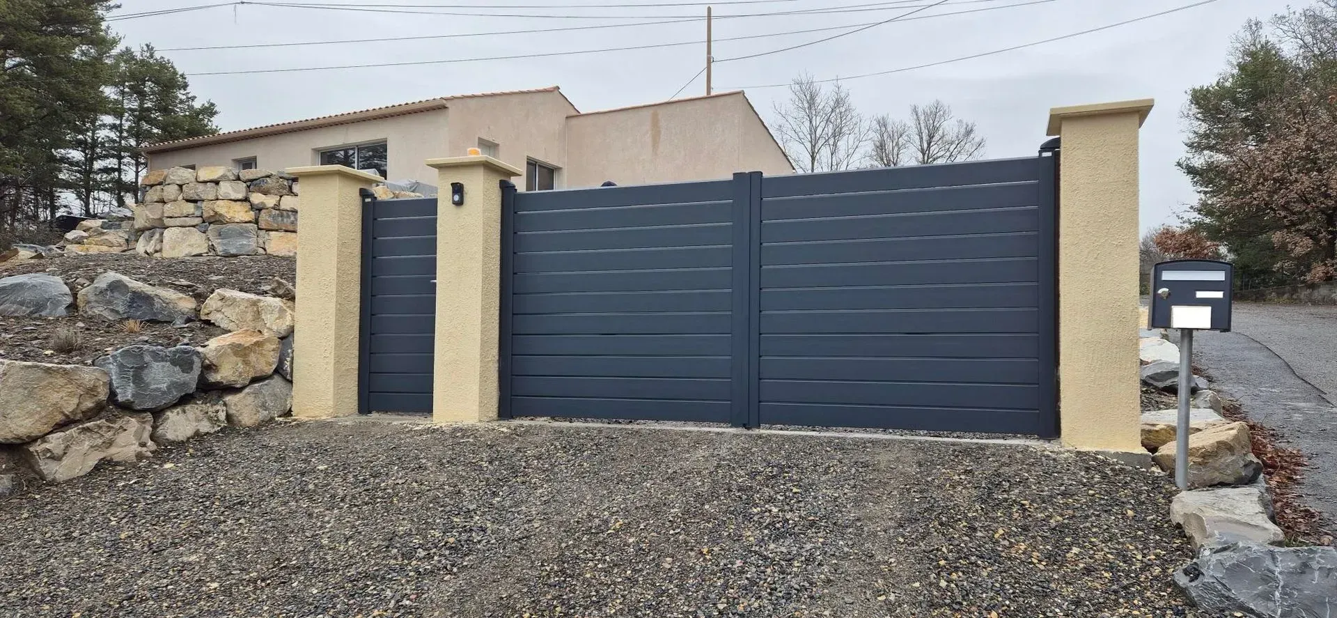 Portail gris foncé avec piliers jaunes et boîte aux lettres ; maison en arrière-plan. Allée de gravier, temps nuageux.