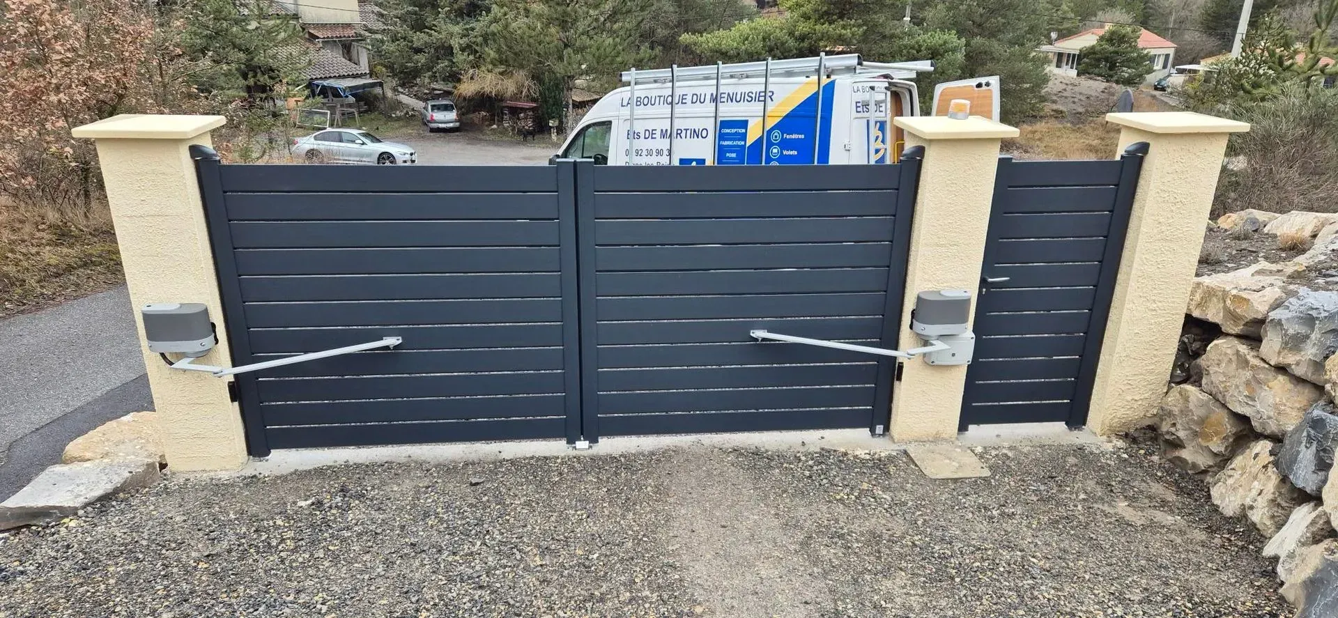 Portail gris automatisé avec deux colonnes en pierre et allée gravillonnée. Une camionnette blanche est visible à l'arrière-plan.