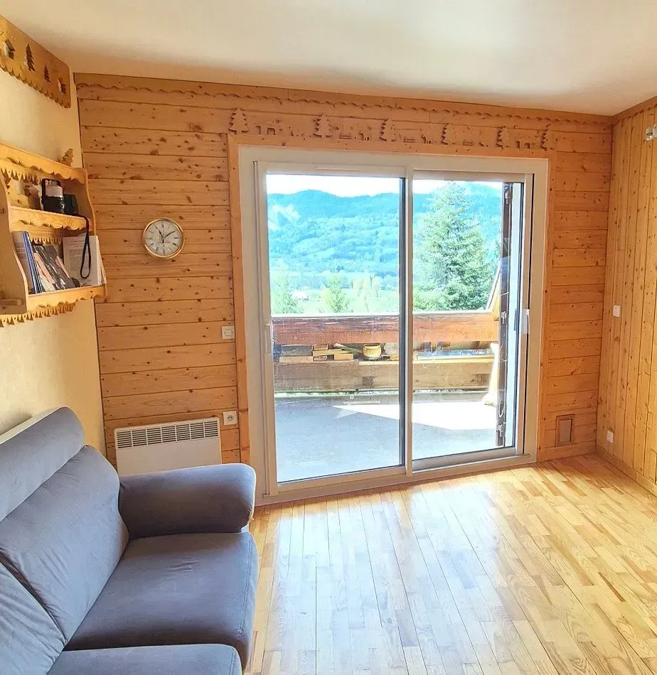 Salon confortable avec lambris, porte-fenêtre coulissante donnant sur un balcon avec vue sur la montagne.