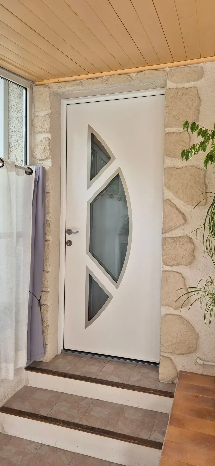 Porte d'entrée blanche avec un motif géométrique en verre, mur en pierre et plafond en bois.