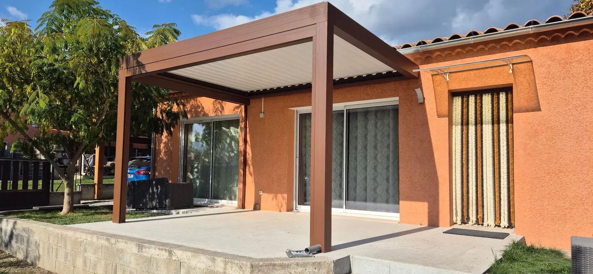 Pergola brune au-dessus d'une terrasse en béton devant une maison aux murs en stuc orange et un arbre.
