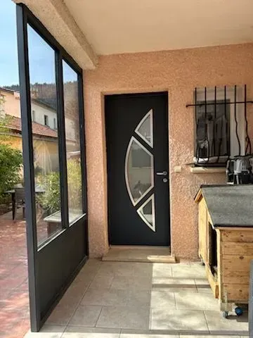 Porte noire ornée de panneaux de verre décoratifs dans une entrée couleur terre cuite, avec un mur vitré sur la gauche.