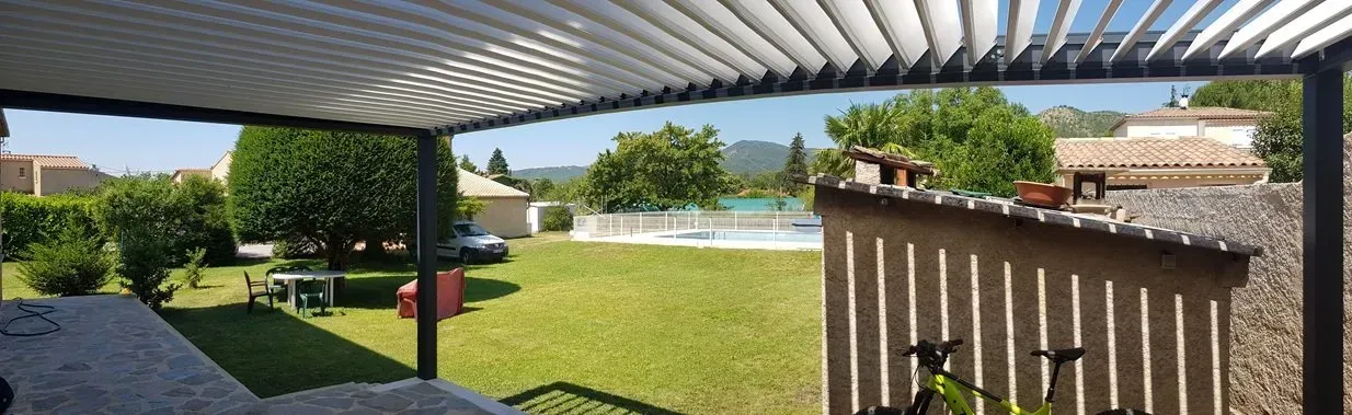 Vue depuis une pergola sur un jardin verdoyant avec des arbres, une piscine et des maisons sous un ciel bleu.