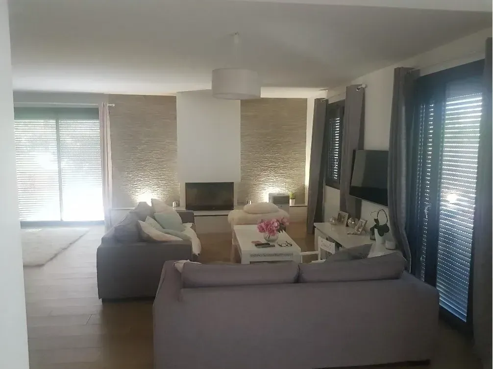 Salon avec canapés gris, mur en pierre, cheminée et fenêtre avec stores.