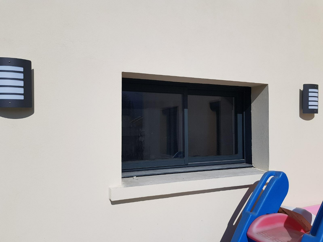 Fenêtre à cadre foncé sur un mur clair, flanquée de deux appliques murales noires ; on aperçoit des équipements de jeux bleus et rouges.
