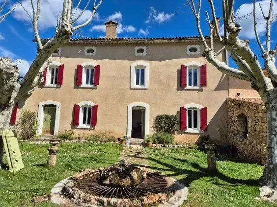 Maison beige à deux étages avec volets rouges et porte verte, encadrée par des arbres, au centre d'une cour herbeuse, sous un ciel bleu.