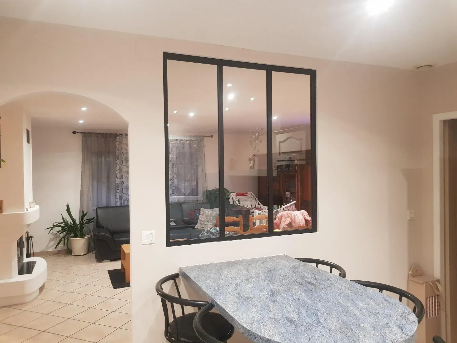 Intérieur avec une fenêtre à cadre noir reflétant un espace de vie, un bar avec deux tabourets et une arche.