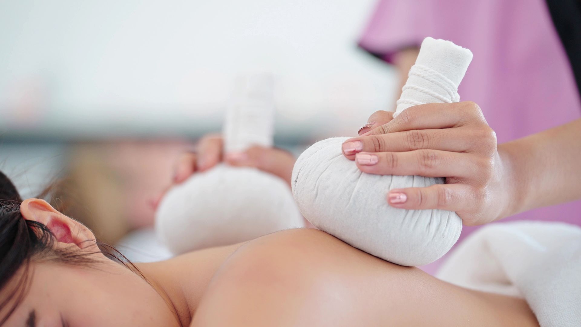 Eine Person erhält eine Spa-Behandlung, bei der ihr von einer Therapeutin in rosa Uniform ein gedämpfter Kräuterkompressen auf den Rücken gelegt wird.
