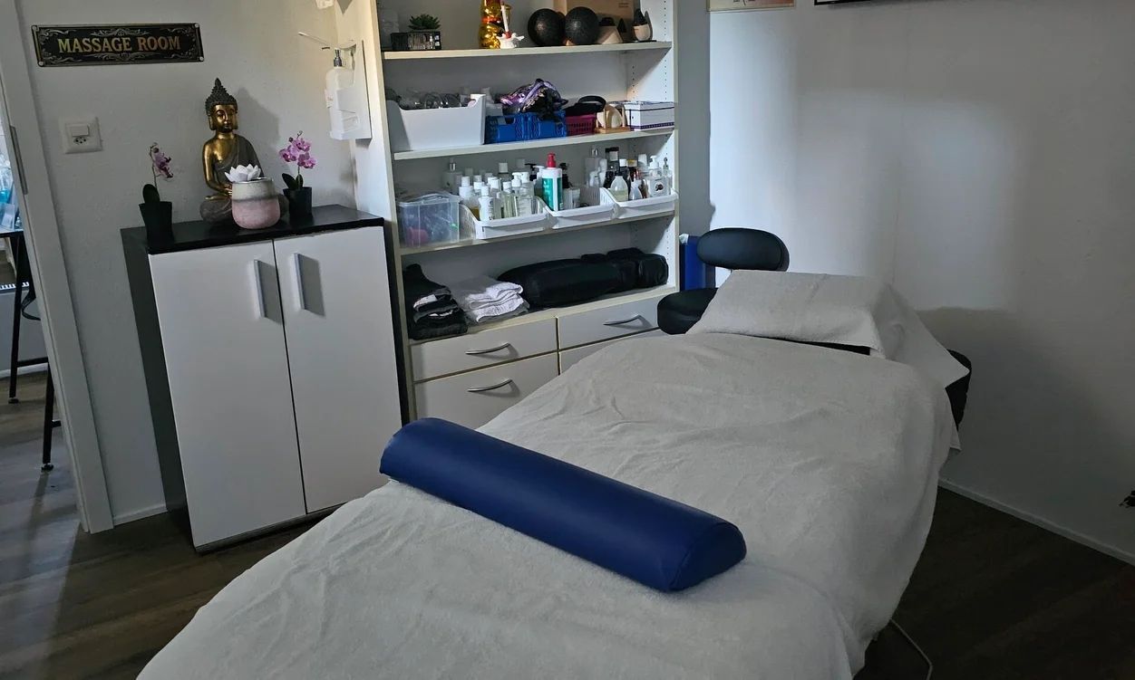 Ein Spa-Behandlungsraum mit einer Massageliege, die mit einem weissen Laken und einem blauen Kissen bezogen ist, neben einem Schrank und Ablageregalen.