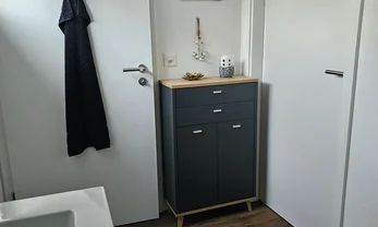 In einem Badezimmer steht ein dunkelgrauer Schrank mit heller Holzplatte vor einer weissen Wand zwischen zwei weissen Türen.