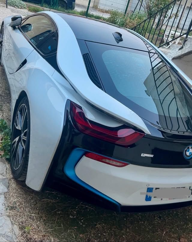 BMW i8 blanche garée en bord de route, vue trois quarts arrière, près d'une clôture et d'herbe.