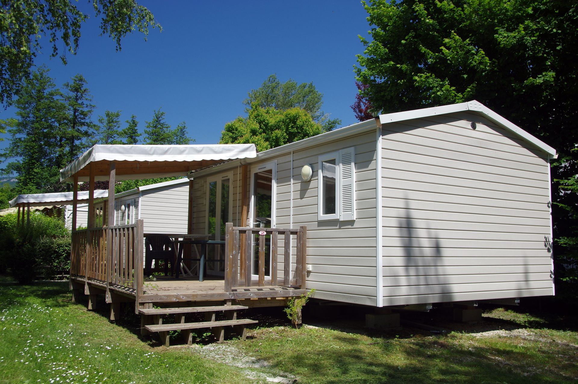 Mobil-home blanc avec véranda et marches dans une zone herbeuse et boisée sous un ciel bleu