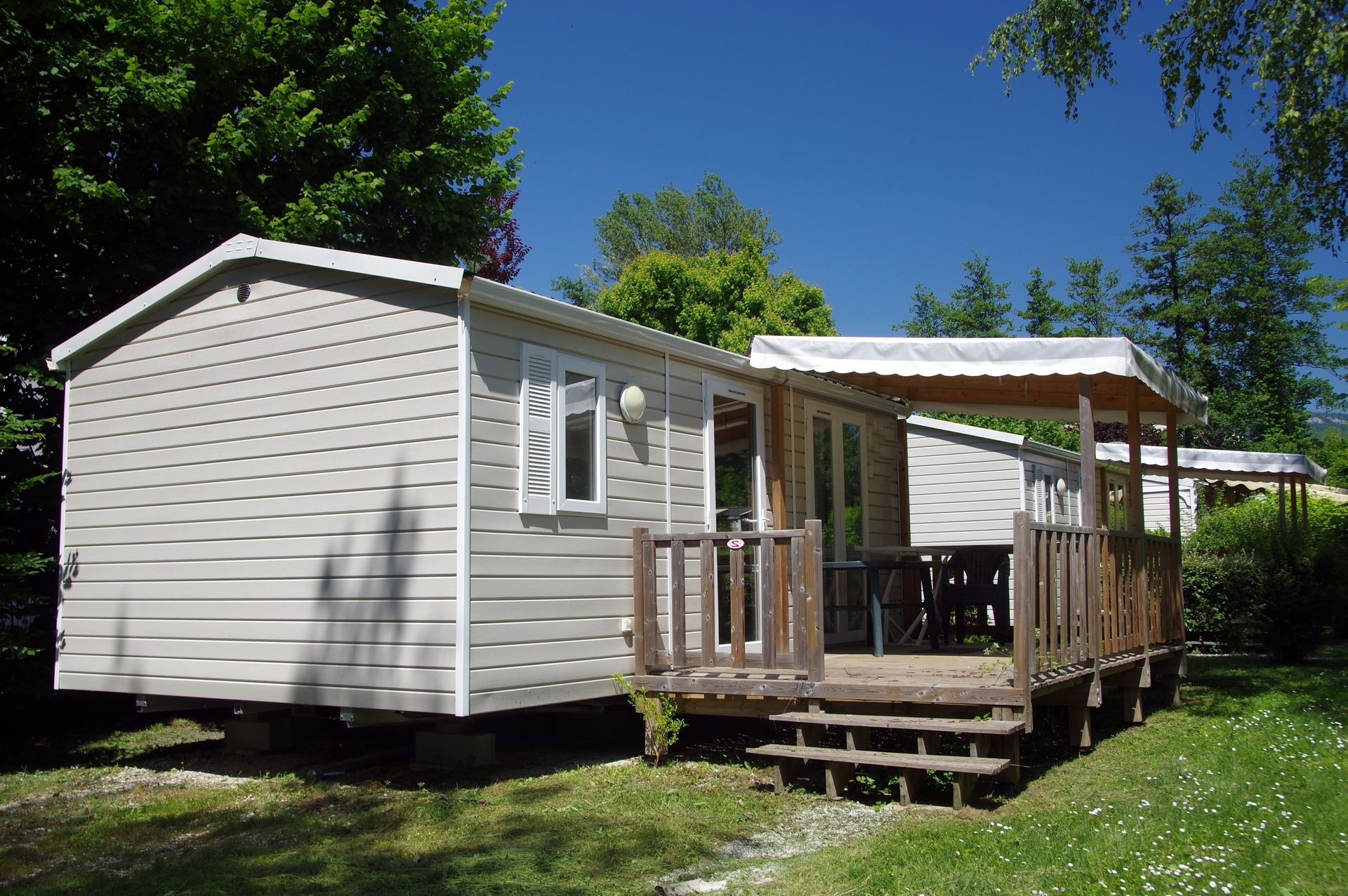Mobil-home blanc avec une petite véranda en bois, entouré d'herbe verte et d'arbres sous un ciel bleu.