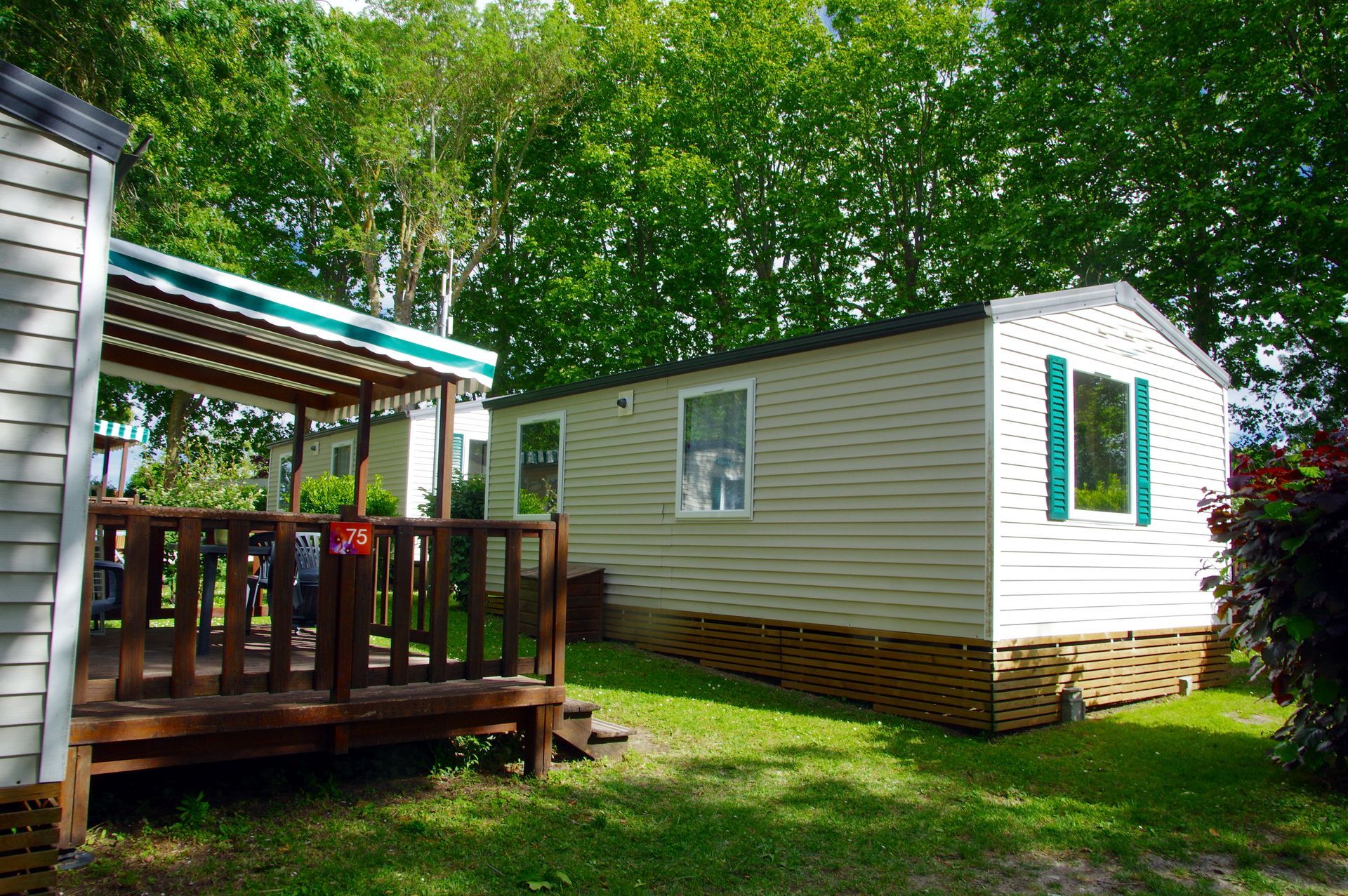 Mobil-home blanc aux volets verts, jouxtant une terrasse en bois sous les arbres, dans une cour herbeuse.