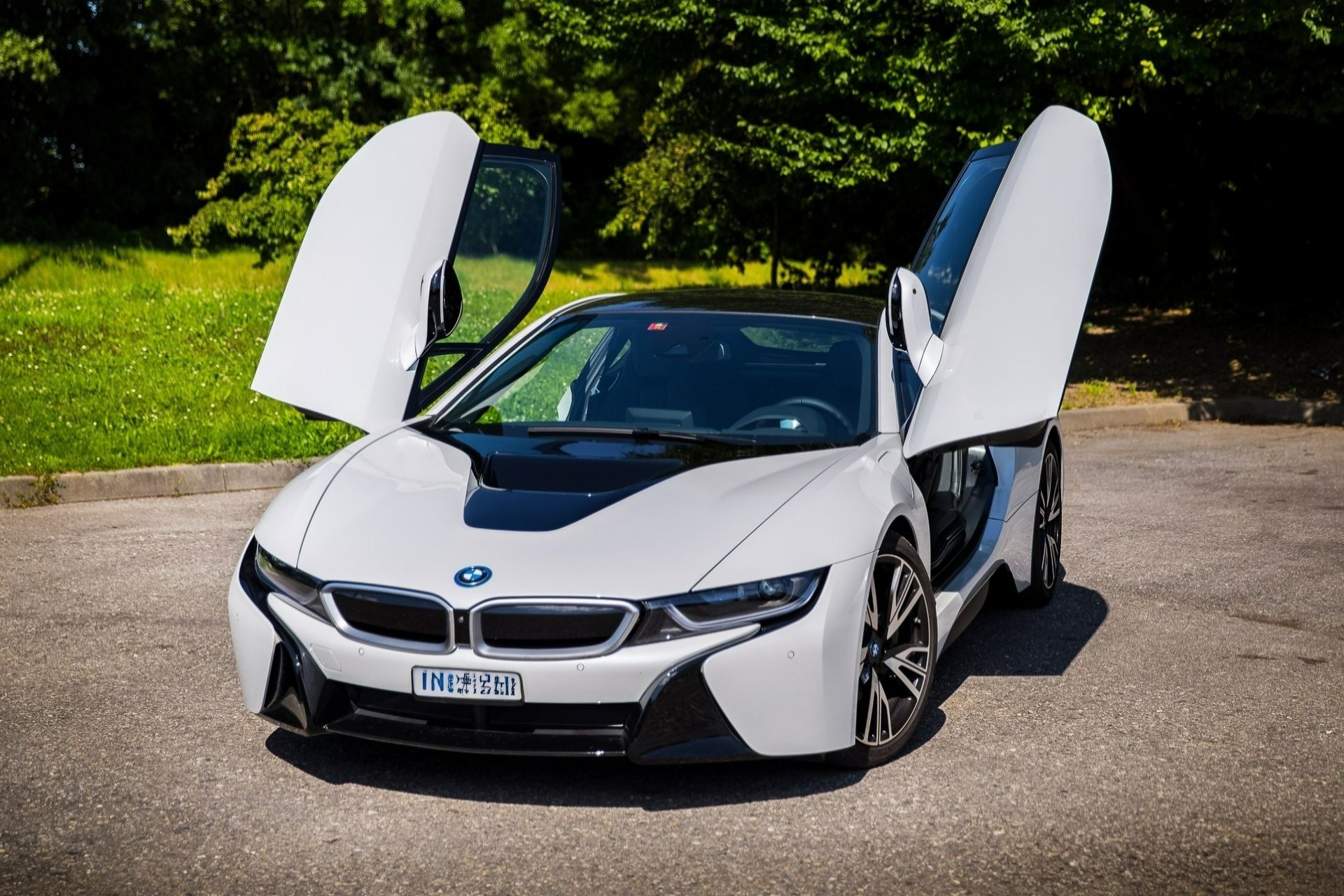 Une BMW i8 blanche, portes papillon ouvertes, est garée sur une route devant des arbres verts.