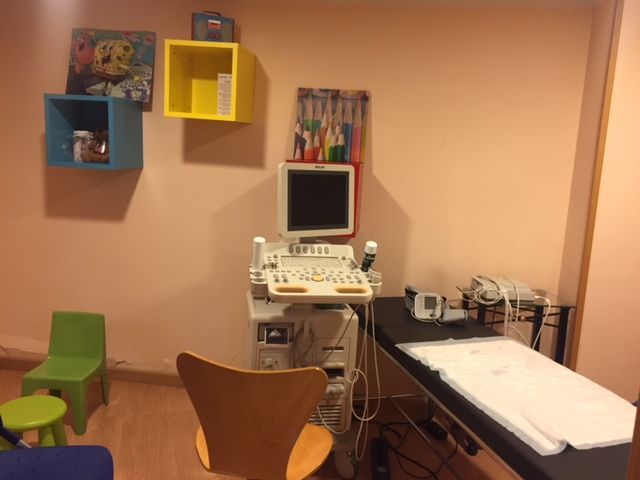 Sala de exploración con equipo de ultrasonido, cama y estanterías de pared de colores.
