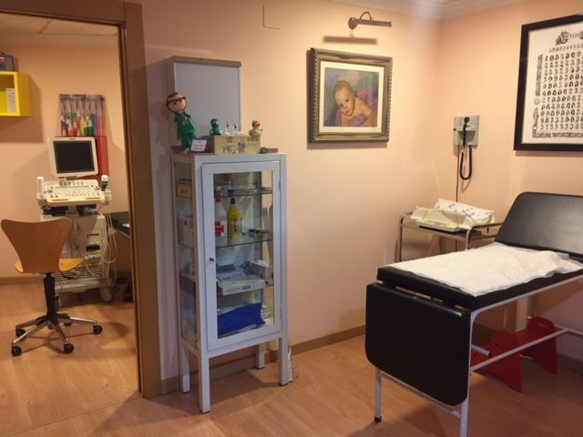 Interior del consultorio médico con equipo médico y mesa de exploración; paredes color durazno, suelos de madera.