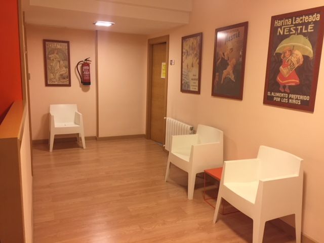 Pasillo con sillas blancas, pósteres y un extintor. Paredes beige y suelo de madera.