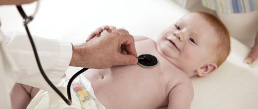 Un médico examina el pecho de un bebé con un estetoscopio. El bebé está recostado sobre una superficie blanca, sonriendo.