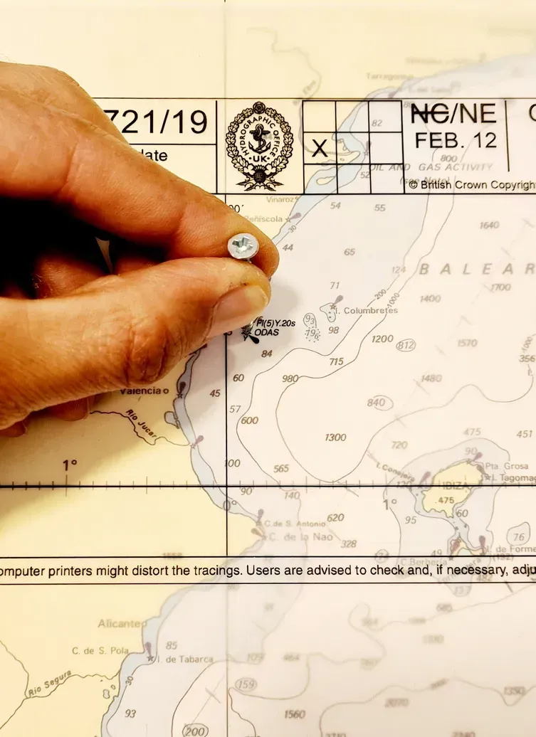Una mano utiliza un alfiler para marcar una ubicación en una carta náutica que muestra curvas de nivel de profundidad y etiquetas geográficas.