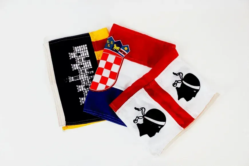Tres banderas superpuestas: la negra y dorada de Flandes, la tricolor croata con su escudo y la bandera de Cerdeña.