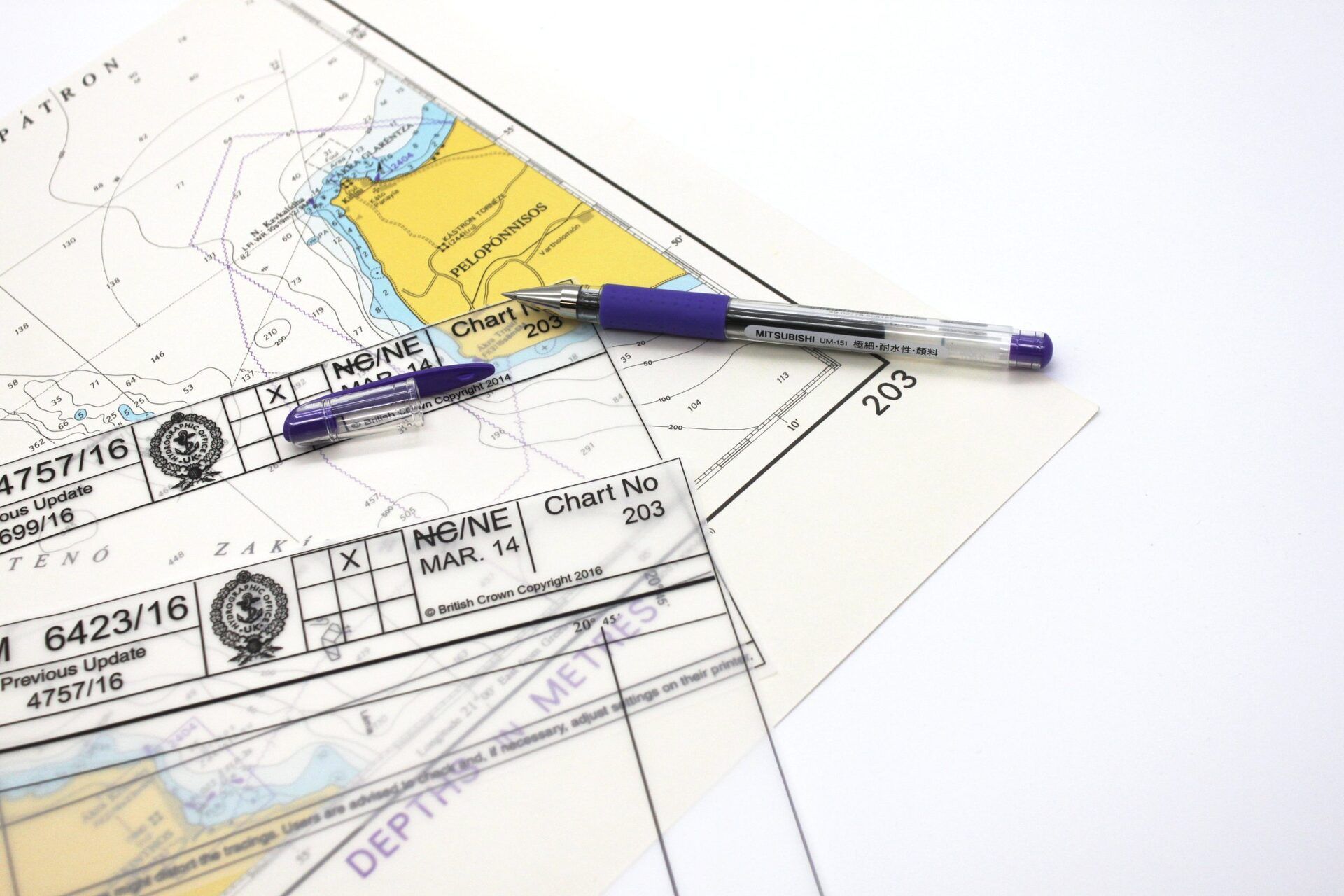 Una carta náutica con una regla paralela transparente apoyada sobre ella, acompañada de dos bolígrafos azules.