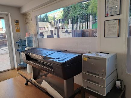 En una oficina con una gran ventana, hay una impresora de gran formato y una impresora láser de varias bandejas sobre mesas.