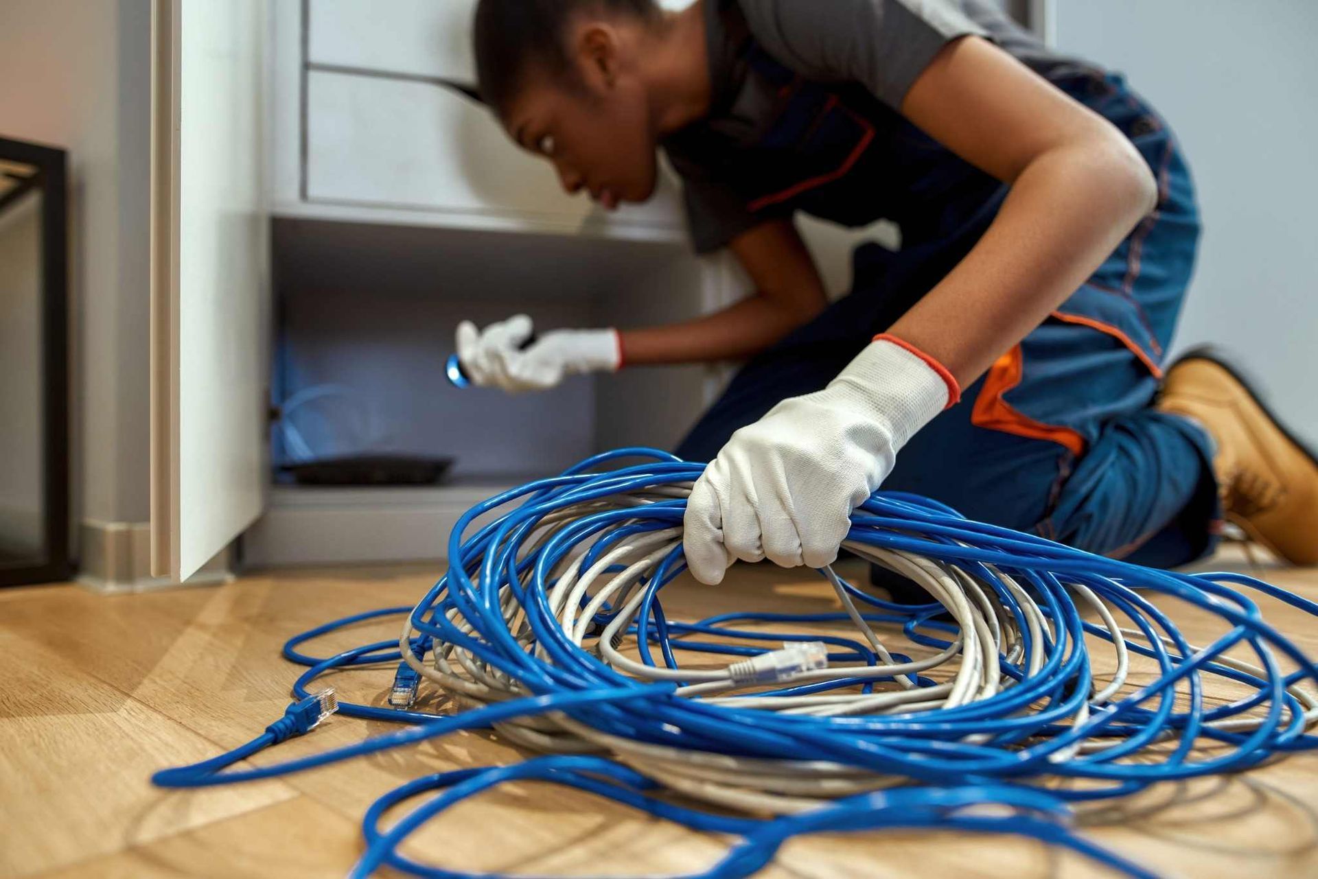 Una persona, arrodillada sobre un suelo de madera, desenreda unos cables azules junto a un armario con las manos enguantadas.