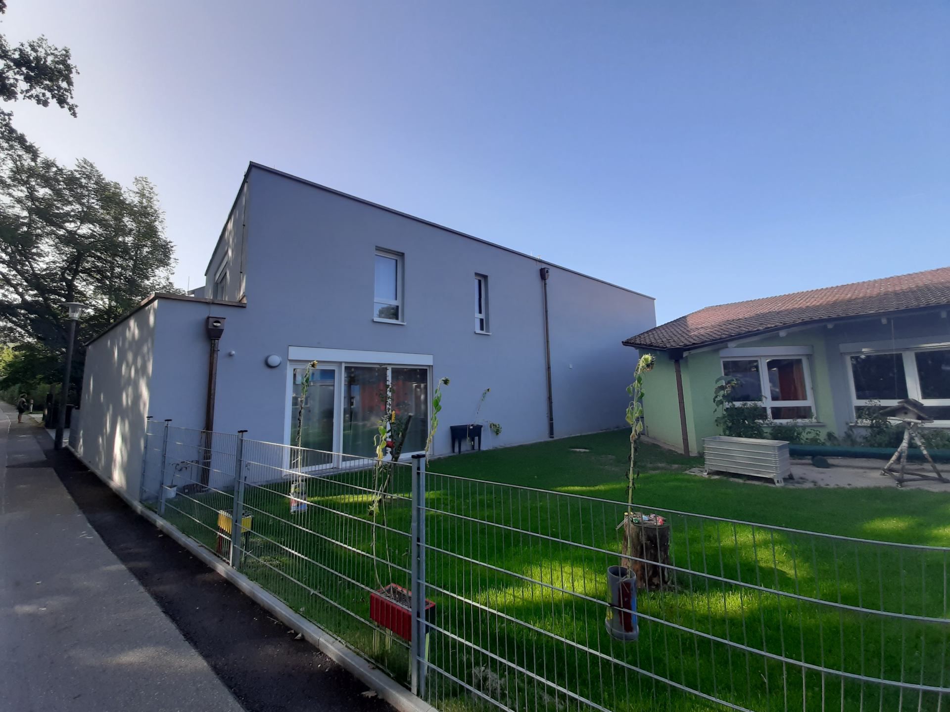 Modernes graues Haus mit rotem Ziegeldach neben einem umzäunten grünen Rasen an einer sonnigen Straße