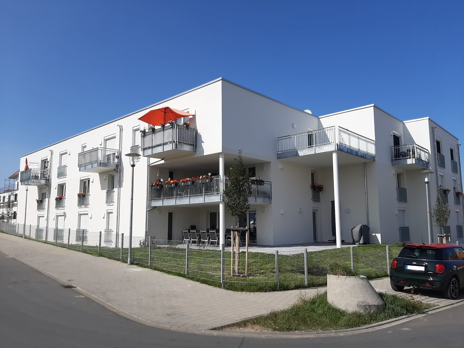 Modernes weißes Wohnhaus mit Balkonen und roten Sonnenschirmen an einer sonnigen Eckstraße